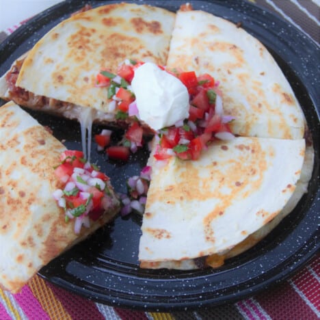 A quarter of a pulled pork quesadilla, topped with pico de gallo and sour cream, is being removed from a black camping plate.