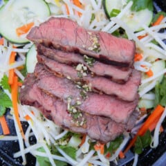 Sliced medium-rare steak is layered on a top of a cucumber, carrot, and bean sprout salad.