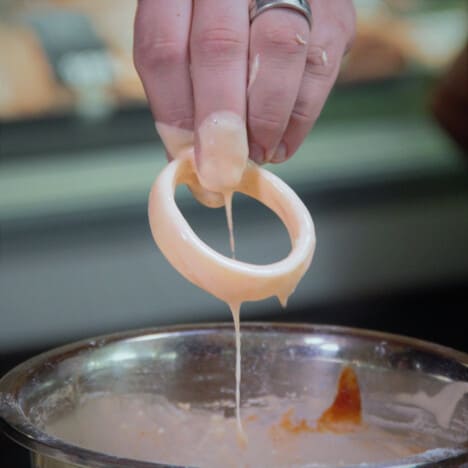 An onion ring being lifted out of the batter with the residual batter running off.