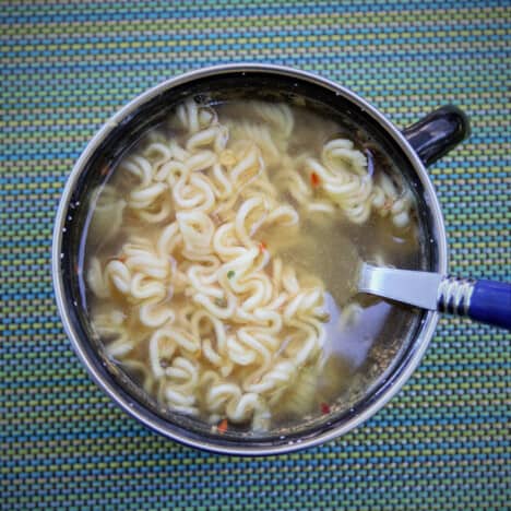 Looking down into a large mug on a blue striped placement with ramen noodles and a fork inside.