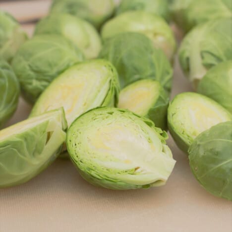 Fresh raw brussels sprouts sitting on a white chopping board, the closest ones are halved.