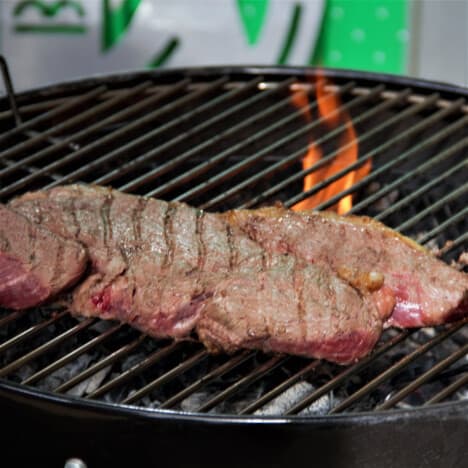 A large rump steak is being grilled over charcoal with a flame flareup.