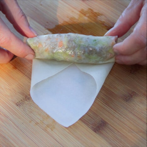 A single spring roll being made in a wrapped on a wooden cutting board.