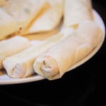 Raw spring rolls line a white serving plate, awaiting to be deep fried.