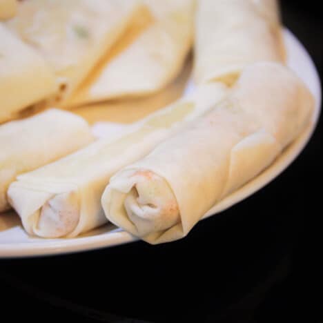 Raw spring rolls line a white serving plate, awaiting to be deep fried.