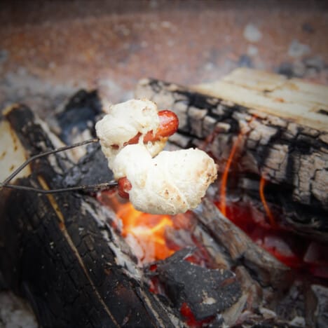 Two mini sausages wrapped in dough are being held over coals with a cooking stick.