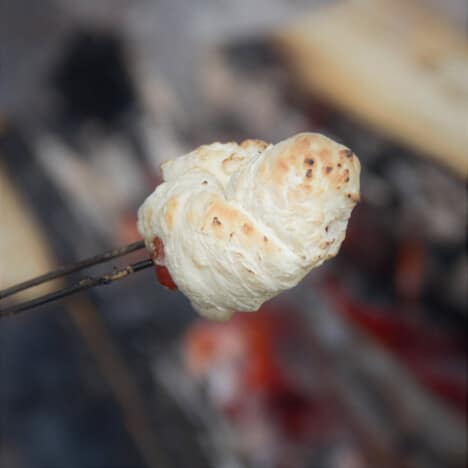 A cooked beef in a blanket is being held on a roasting stick over a campfire.