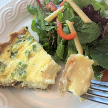 A slice of bacon and onion quiche sits on a white plate next to a green salad.
