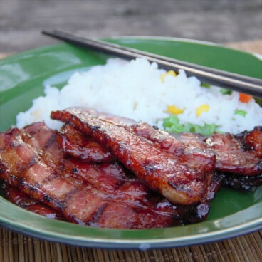 Grilled Asian pork belly served is served on a green camping plate with a side of rice and chopsticks.