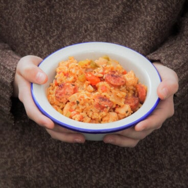 A person holding with both hands a full camp bowl of jambalaya with their knitted pull-over in the background.