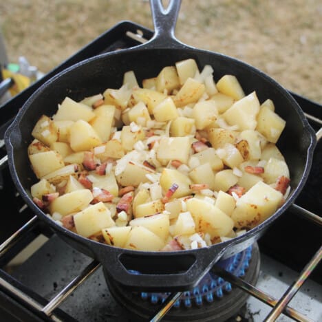 Breakfast potatoes cooking in a skillet over a camp gas stove.