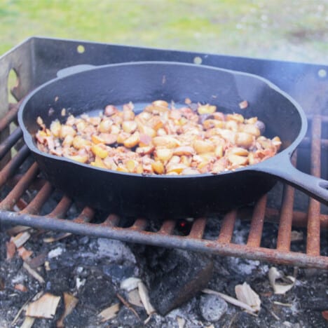 Breakfast potatoes cooking in a skillet over a campsite fireplace.