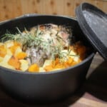 A Dutch oven has its lid to the side exposing a perfectly cooked roast lamb surrounded by roast vegetables.