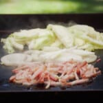 Bacon, onions, and cabbage being cooked on a flat top grill.