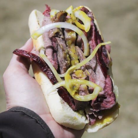 A hand is holding a finished ribeye sandwich with meat, onions, and mustard.