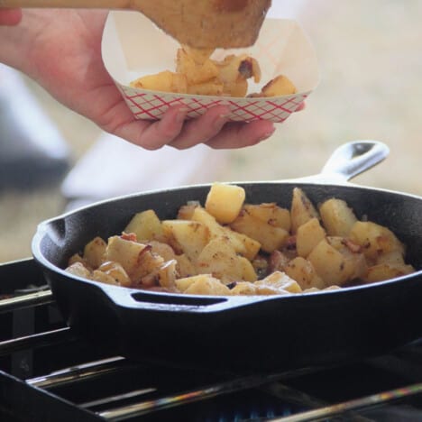 Breakfast potatoes being served into a small paper dish from a skillet sitting on a camp stove.