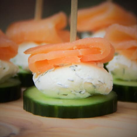 A close up of a smoked salmon cucumber bite with a layer of cucumber, herbed creamed cheese, and smoked salmon held together with a toothpick.