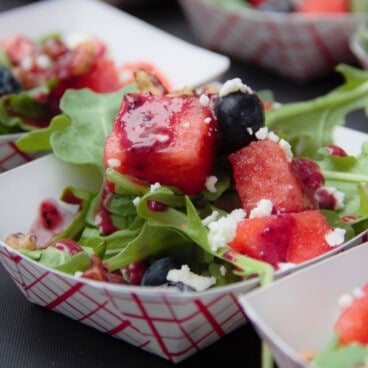 A close up shot of a watermelon and blueberry salad drizzled with sauce, served in a red-checkered paper boat.