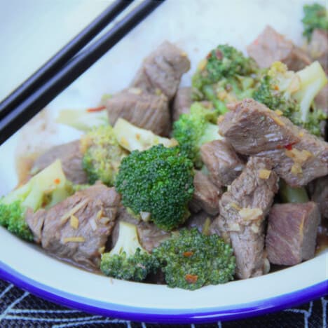 Chunks of beef and broccoli florets are cooked in a sauce and served over rice with chopsticks.