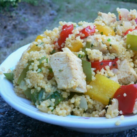 A closeup of a white plate with chicken and bell pepper couscous, with multiple colors of bell peppers.