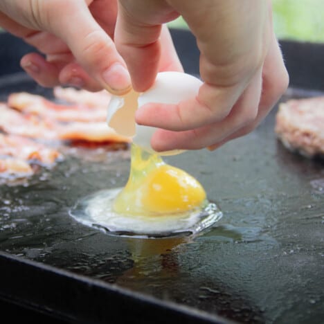 An egg being cracked onto a flat-top grill where bacon and the sausage patty are already cooking.