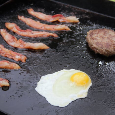 A flat-top grill with bacon, egg, and sausage patty cooking.