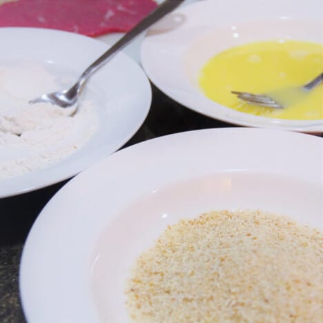 Three white shallow dishes with the three crumbing station components in them, flour, eggs, and breadcrumbs.
