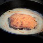 Schnitzel sizzling in a skillet with the top side golden brown after having done its first turn.