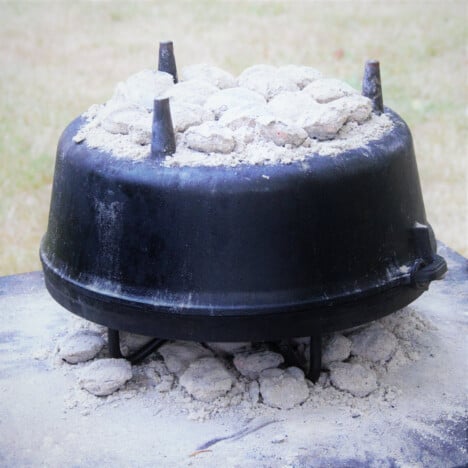 A Dutch oven is upside down with coals on top and underneath while it is cooking.