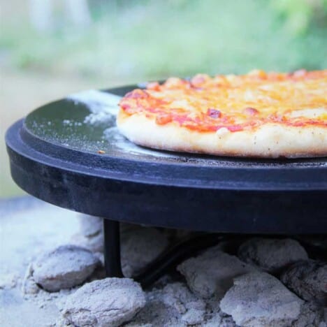 A ham pizza is sitting on a Dutch oven lid over coals after being cooked, awaiting to be moved and served.
