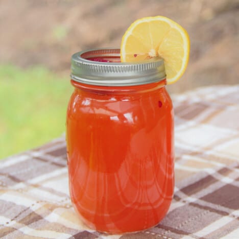 A Raspberry Arnold Palmer is in a canning jar garnished with a slice of lemon, sitting on a brown checkered tablecloth.