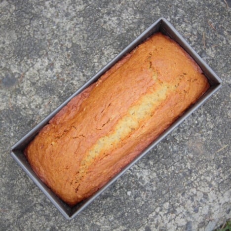 A golden brown loaf of banana bread sits in the baking tin on a concrete background.