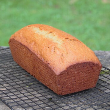 A golden brown loaf of protein banana bread rests on a cooling rack with a grassy background.