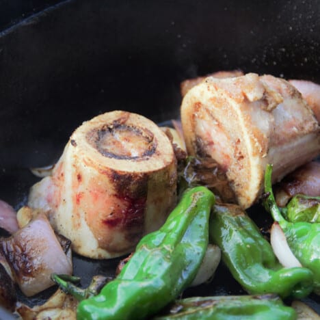 Two rings of bone marrow are in a skillet, surrounded by shishito peppers and red onions.