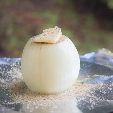 A raw onion with a pat of butter and herb seasoning sits on a piece of foil.