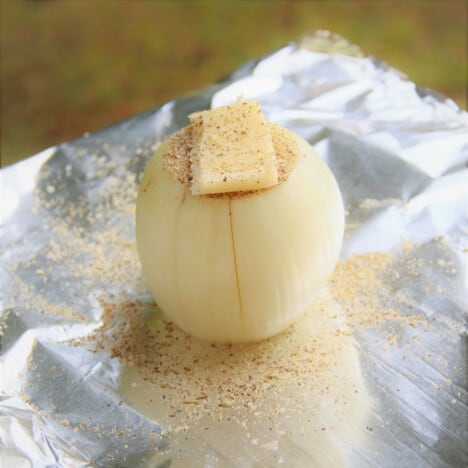 Looking across a raw onion with a pat of butter and herb seasoning, resting on a piece of foil.