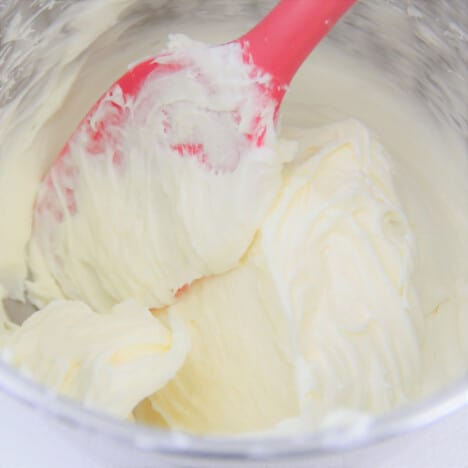 A red spatula stirs the cream cheese frosting in a stainless steel bowl.
