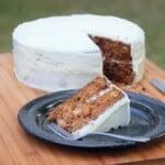 A carrot and walnut cake sitting on a wooden cutting board with one slice removed and placed on a black camping plate.