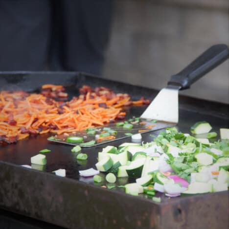 Piles of carrots and bacon and onions and peppers cook on a flat top griddle.