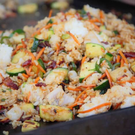 Looking down onto a pile of griddle fried rice with zucchini, shredded carrots, and pieces of bacon.