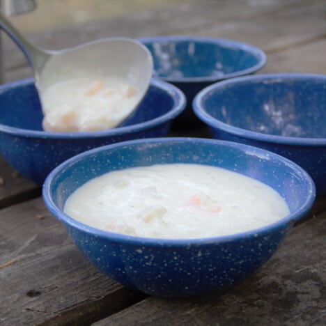 The quick shrimp chowder is being ladled into four blue camping bowls on a picnic table.