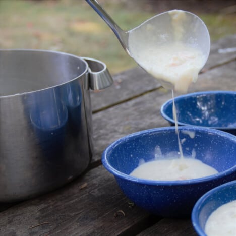 Quick shrimp chowder is being ladled from a large stock pot into three blue camping bowls.