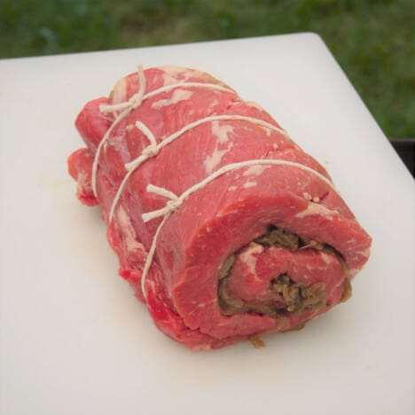 A raw sage and onion beef roulade tied with twine, sitting on a white cutting board.