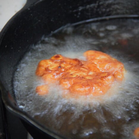 A single golden-brown corn fritter is frying in a cast iron skillet full of oil.