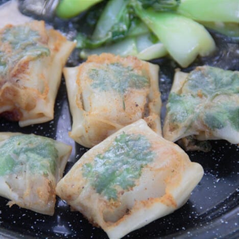 Several golden brown packets of cooked Asian Rice Paper Fish resting on a platter in front of green vegetables.