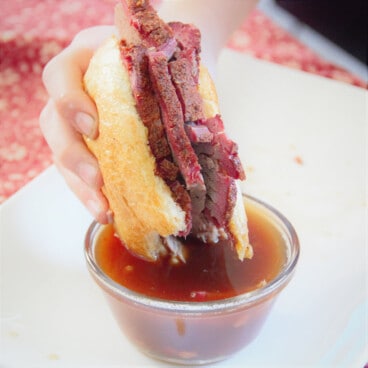 A roll with slices of brisket being dipped into a BBQ brisket au jus.