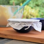 A blue camp bowl with a prepared steamed pudding topped with baking paper and tied with butchers twine. The mixing bowl with the remaining batter is in the background.