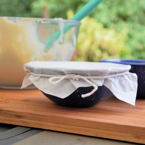 A blue camp bowl with a prepared steamed pudding topped with baking paper and tied with butchers twine. The mixing bowl with the remaining batter is in the background.