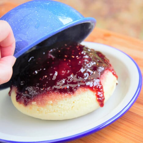 The blue camp bowl being removed from the pudding, sitting upside down on a camp plate, exposing the raspberry jam running down the side of the pudding.