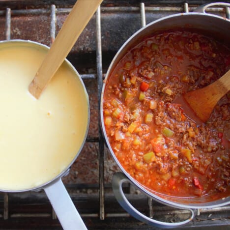 Looking down on a saucepan of béchamel sauce and bolognese sauce cooking on a camp gas stove.